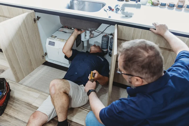 Annual Plumbing Inspection, Two plumbers inspecting under-sink pipes in modern kitchen during routine home checkup.