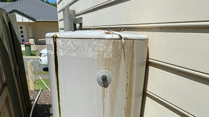 Leaking outdoor hot water system with rust and water stains on tank, showing common hot water problems that need professional plumbing repairs.