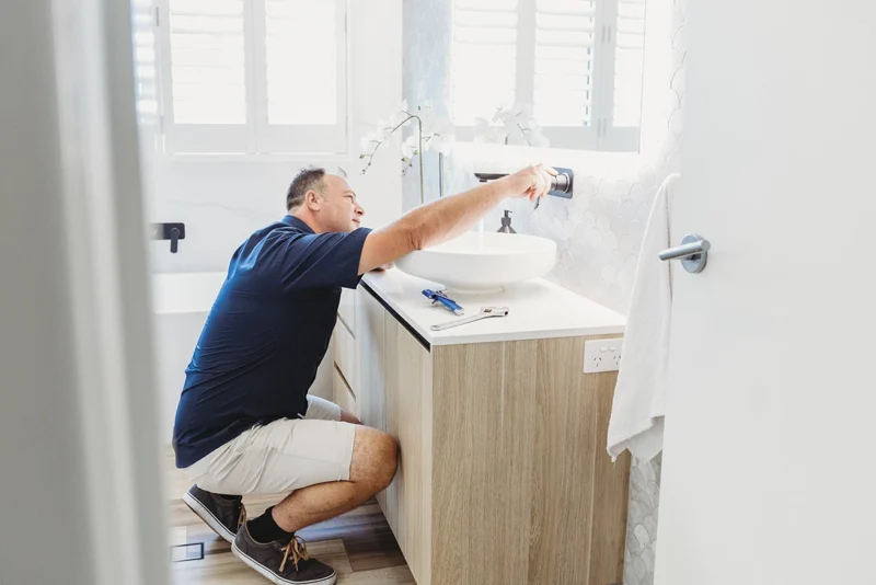A professional plumber from Assure Fix Plumbing performing preventative plumbing maintenance in an Ipswich bathroom, inspecting pipes under the sink.