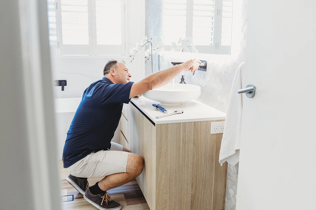 A professional plumber from Assure Fix Plumbing performing preventative plumbing maintenance in an Ipswich bathroom, inspecting pipes under the sink.