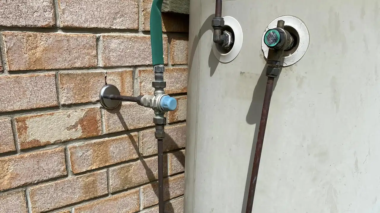 Silver tempering valve with blue cap on the hot water pipe beside an electric hot water system